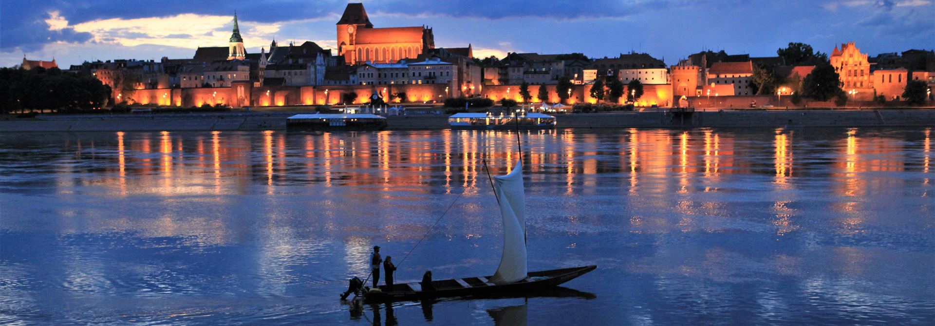 na tle rozświetlonej, wieczornej panoramy Torunia płynie tradycyjna łódź na żaglu z trzema osobami na pokładzie. Zdjęcie pokazuje rejsy w Toruniu.